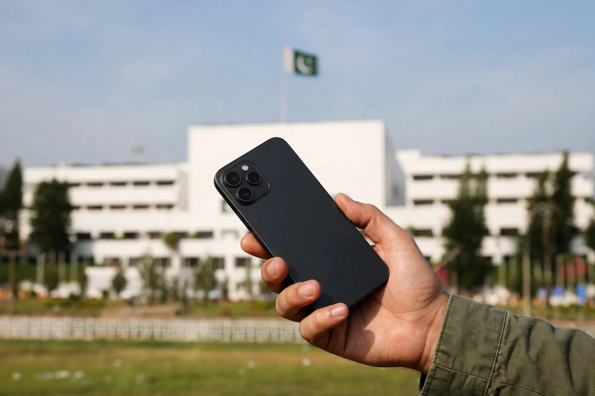 A hand holding a black smartphone in front of a white government building with a Pakistani flag flying above it